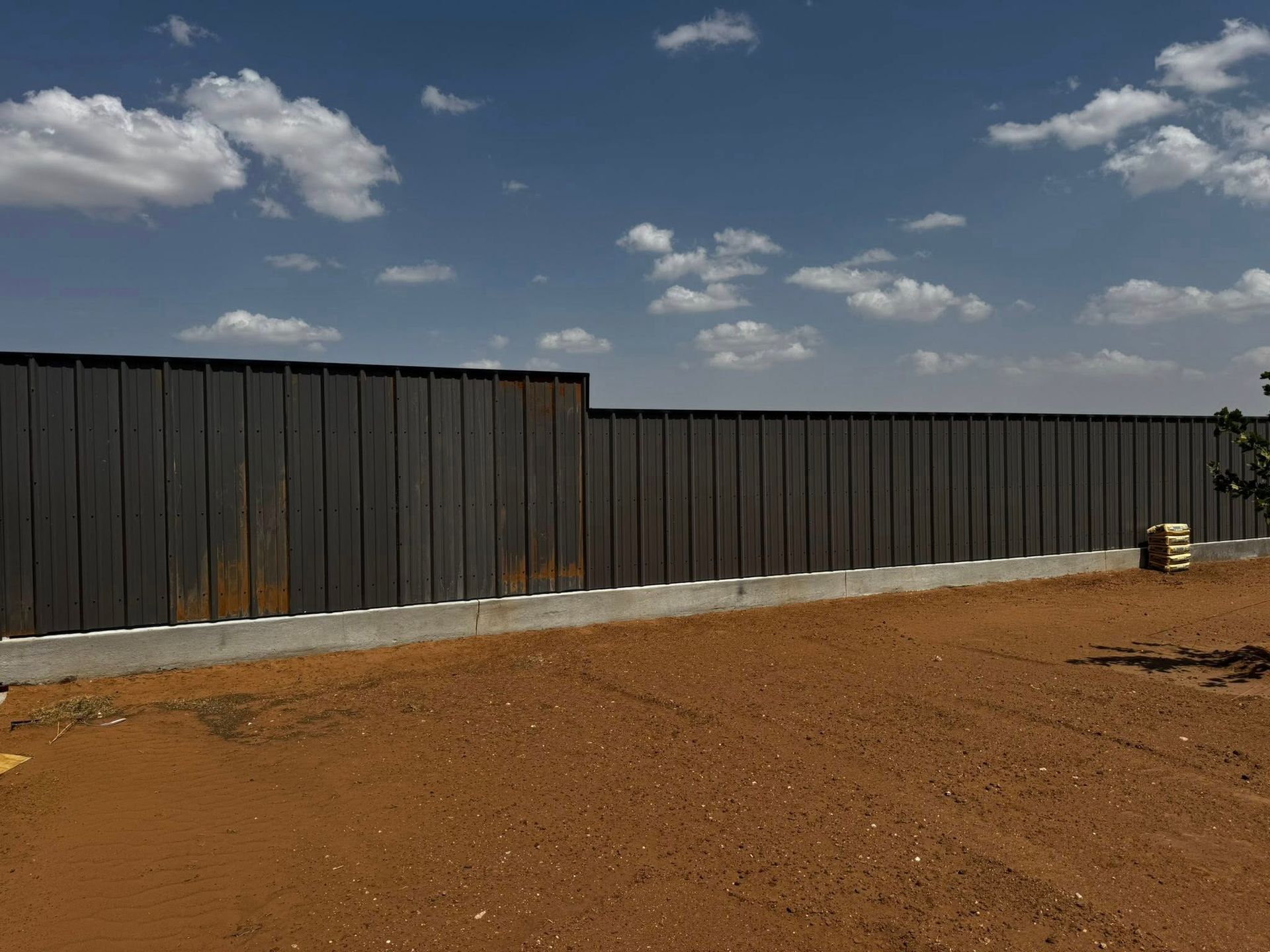Dark metal fence on a concrete base, against a blue sky with clouds, in a dirt yard.
