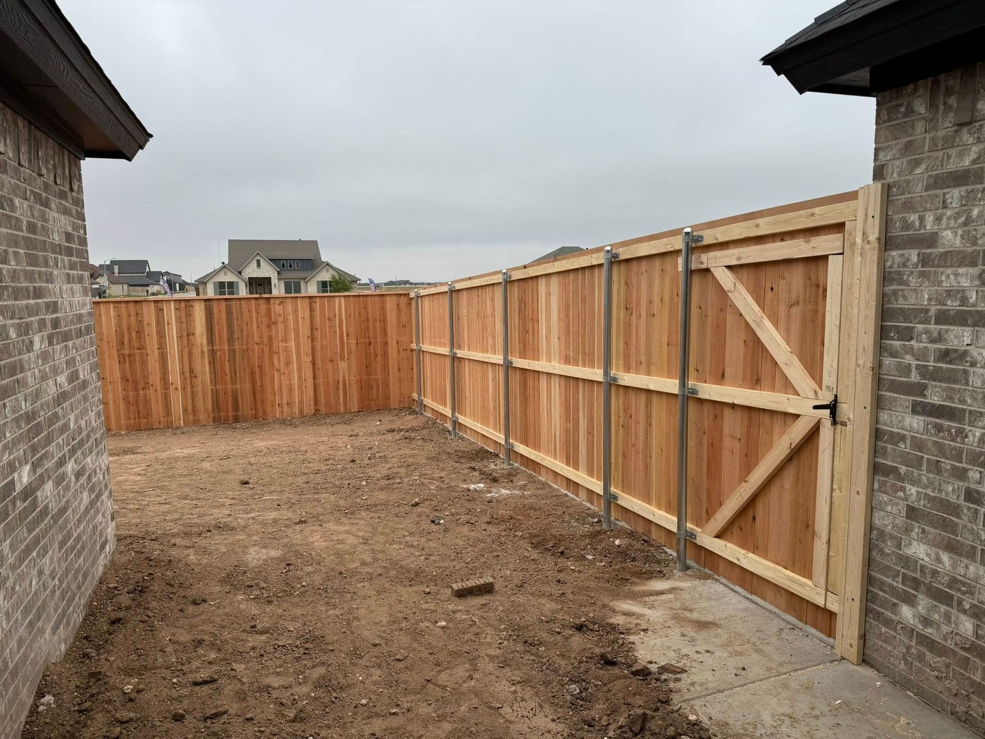 Wooden fence and gate enclose a backyard between brick houses on an overcast day.