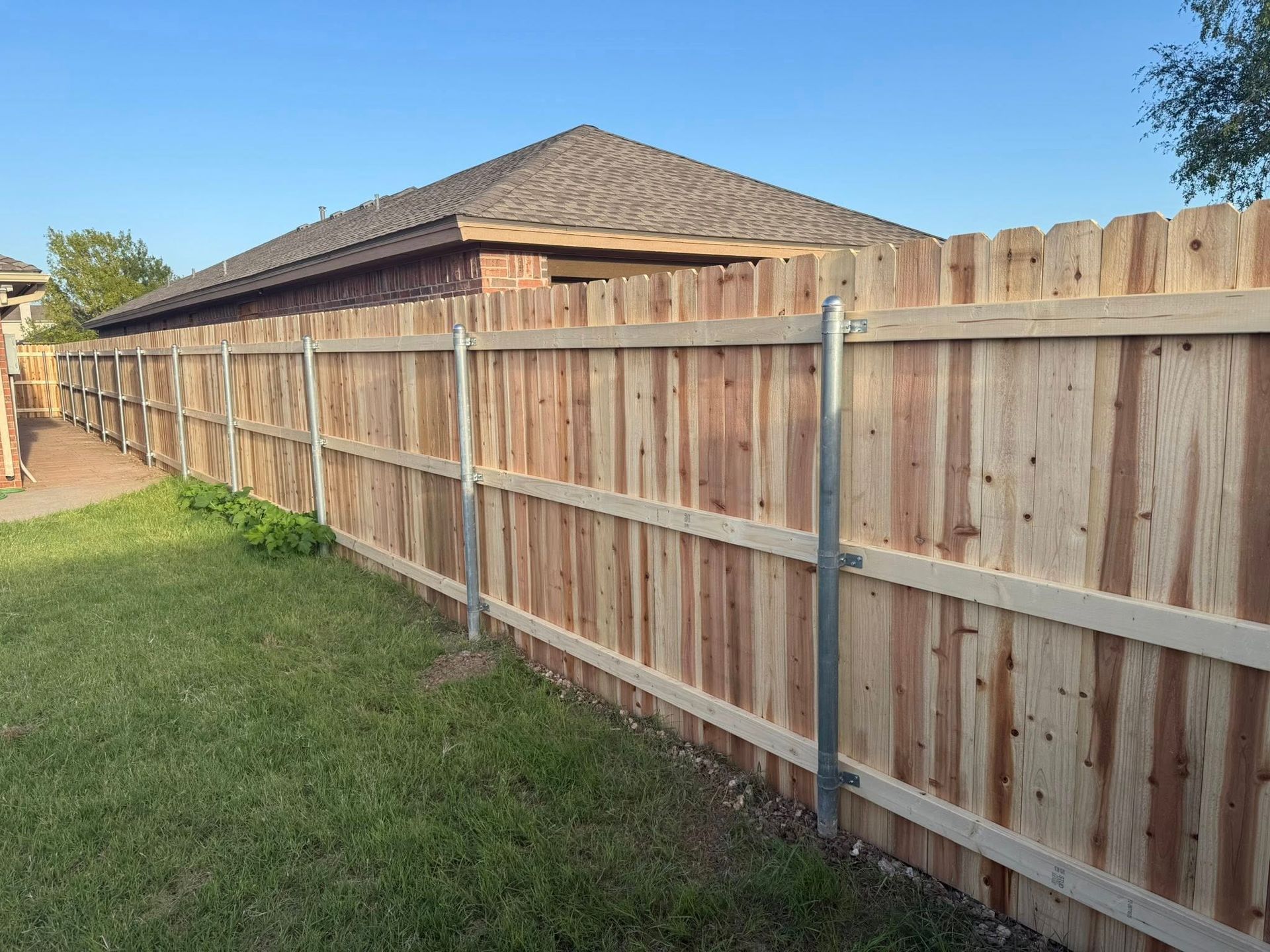 Wooden fence bordering a green lawn, with a house and blue sky in the background.