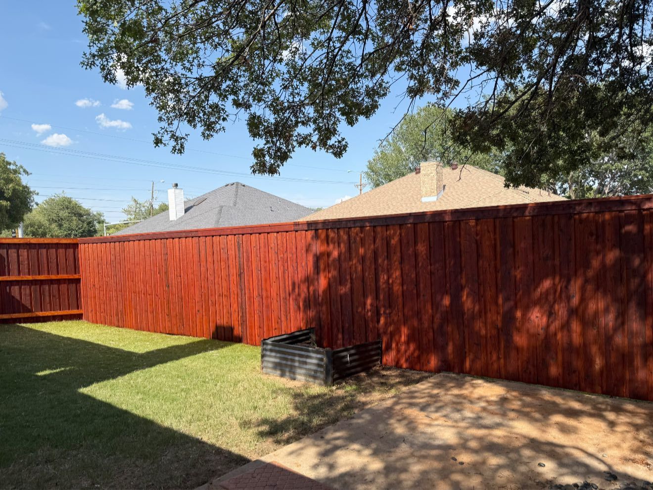 Red stained wooden fence in a backyard with green grass and trees under a blue sky.