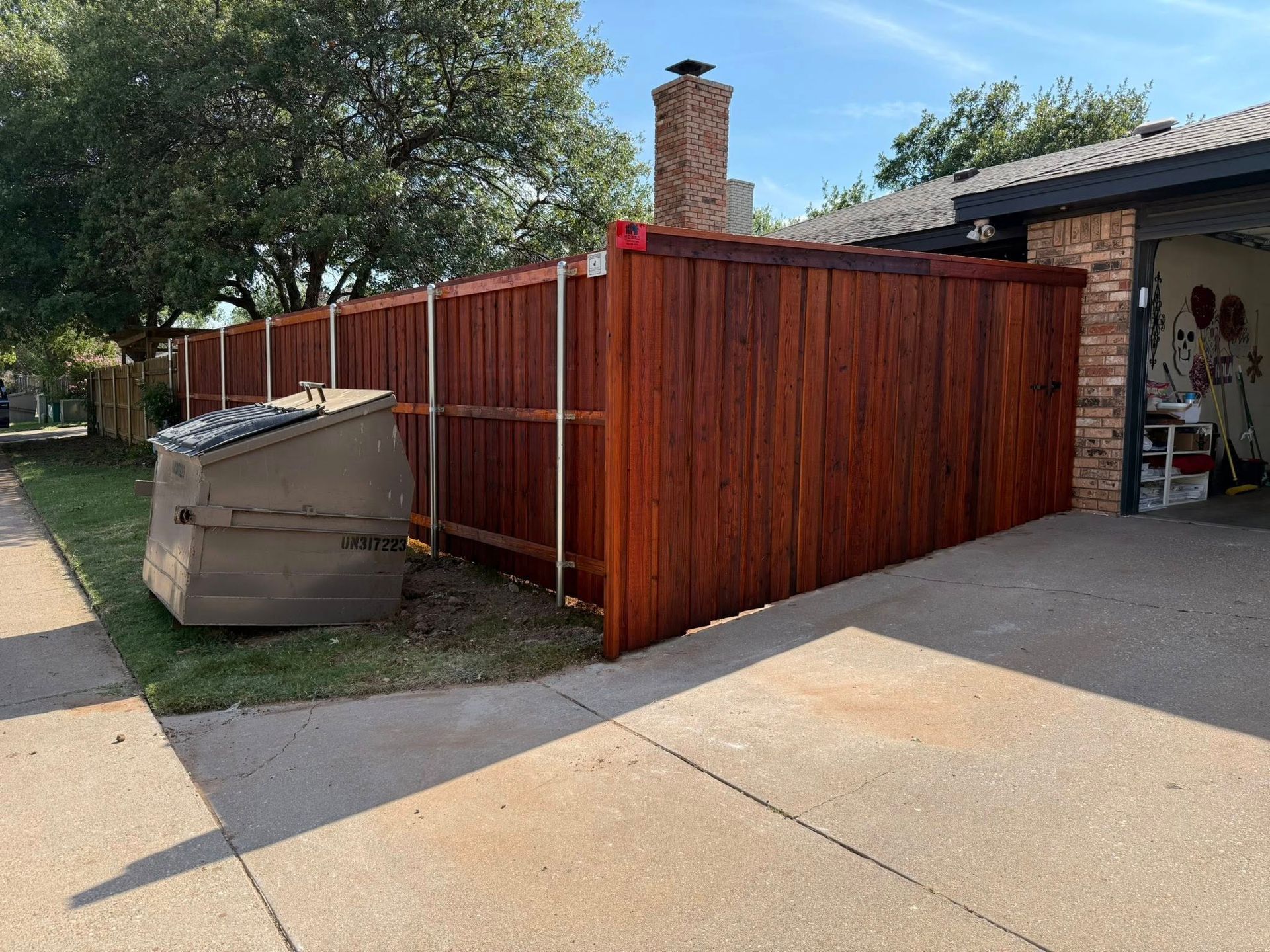 Brown wooden fence beside a driveway and garage on a sunny day. A large, brown utility box sits in the grass.
