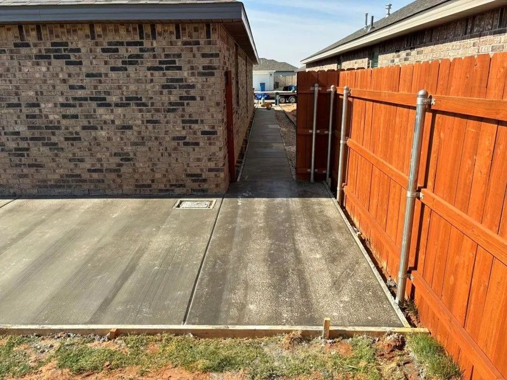 Concrete pathway between a red-brick building and a stained wooden fence. A gate is visible at the end of the pathway.