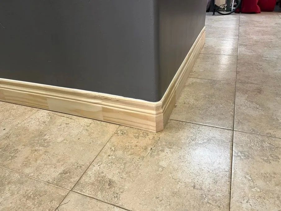 Close-up of a corner with a gray wall, light-colored wood molding, and tan tile flooring.