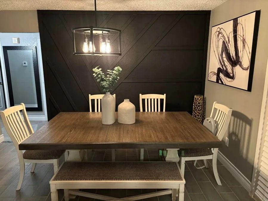 Dining room with a black accent wall featuring a geometric design, a wooden table, and white chairs with bench seating. Artwork and a chandelier are present.