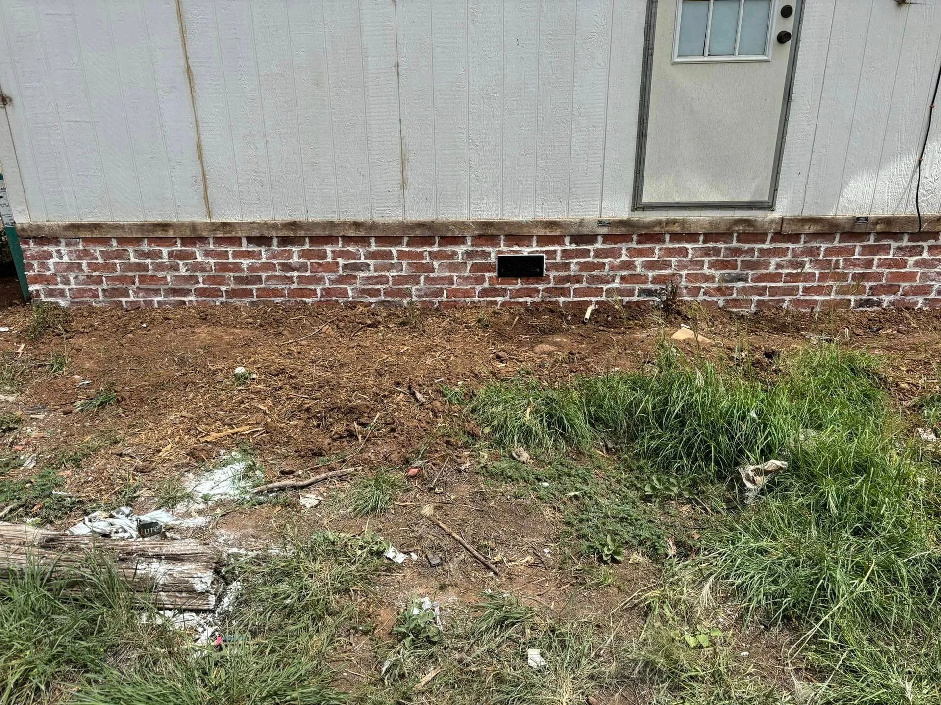 A section of a building with a red brick foundation, a beige wall, and a white door. Dried grass and weeds cover the ground in front.