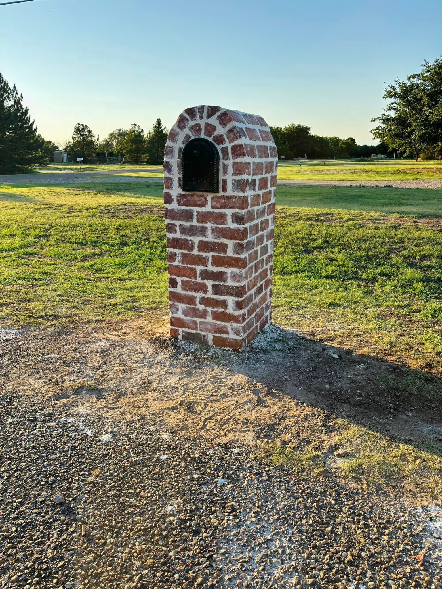 Brick mailbox with arched top, set on gravel driveway, surrounded by grass and trees under a clear sky.
