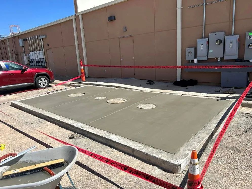 Newly poured concrete slab with three manhole covers, cordoned off with red tape, next to a beige building. A red car is parked nearby.