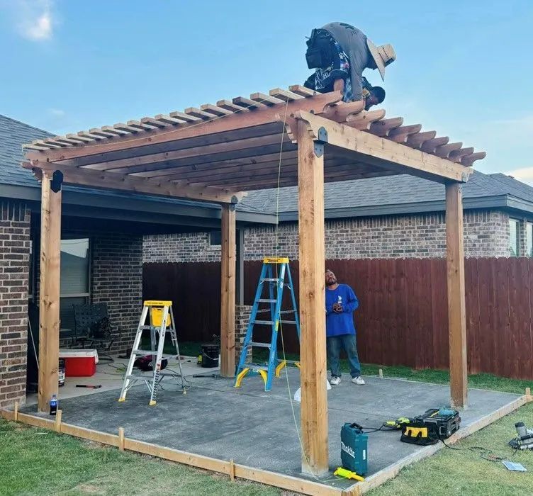 Construction of a wooden pergola in a backyard. Workers are on ladders, building on a concrete patio next to a house and fence.