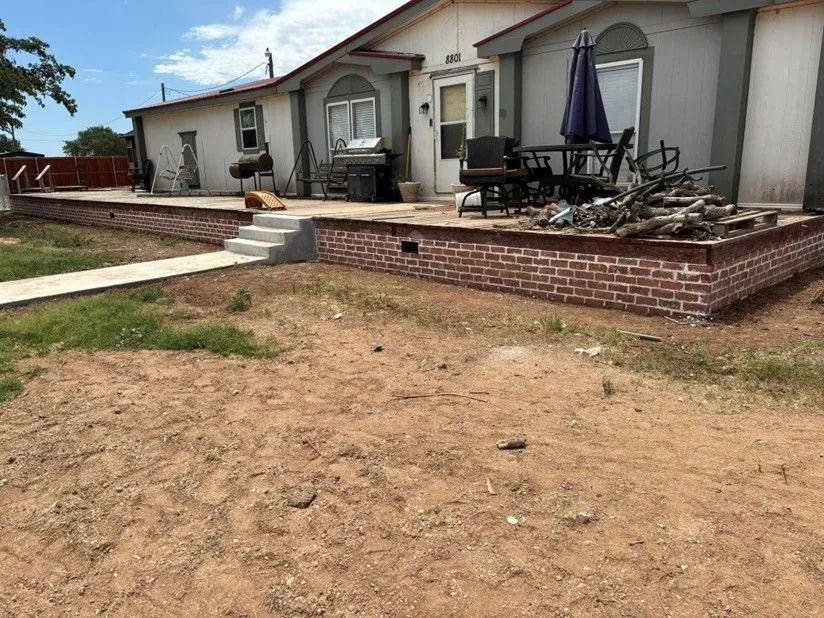 A light-colored house with a brick-lined porch has a patio set and debris. A sidewalk leads to the steps.