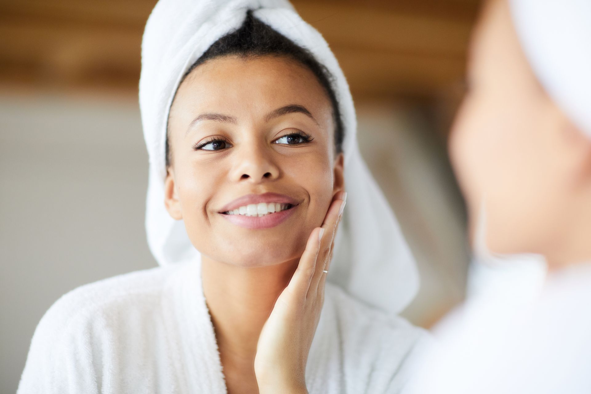 Woman in bathrobe and towel touching face, smiling in mirror.