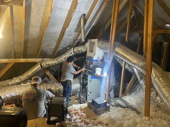 Three people working on an HVAC system in an attic. Ductwork, insulation, and tools are visible.
