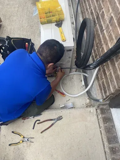 Person in blue shirt repairing an AC unit outside a brick building. Tools and pipes are present.