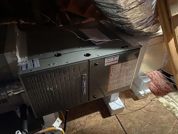 HVAC unit in an attic setting. Gray and black unit with ductwork, insulation, and wooden supports.