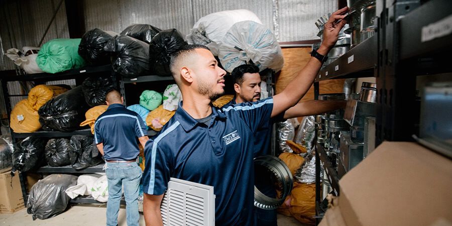 Two men in a warehouse, one reaching for shelf items while another looks on. Bags of material are stacked nearby.