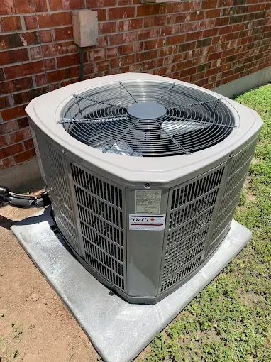 An outdoor air conditioning unit on a concrete pad near a brick wall and grass.