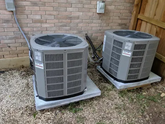 Two air conditioning units on concrete pads beside a brick wall and a wooden fence.