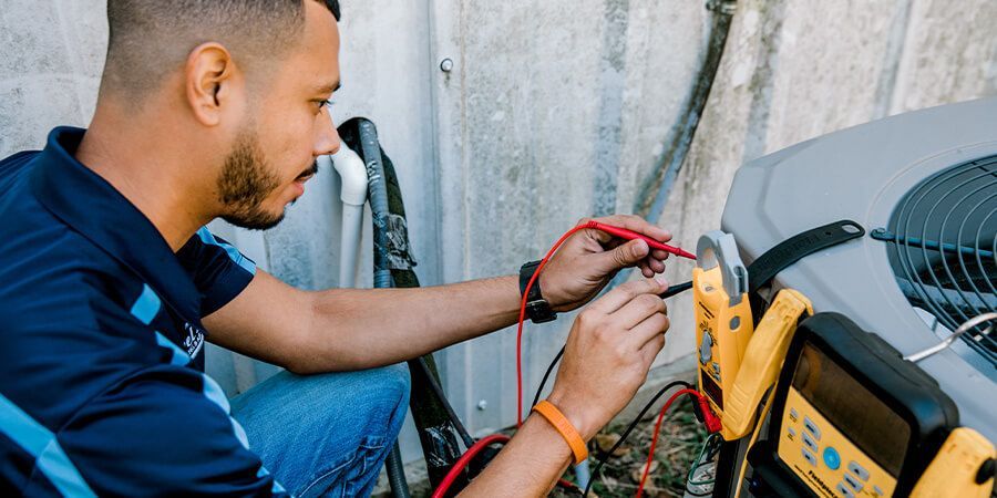 An HVAC technician kneels, using a multimeter to test an air conditioning unit outside.