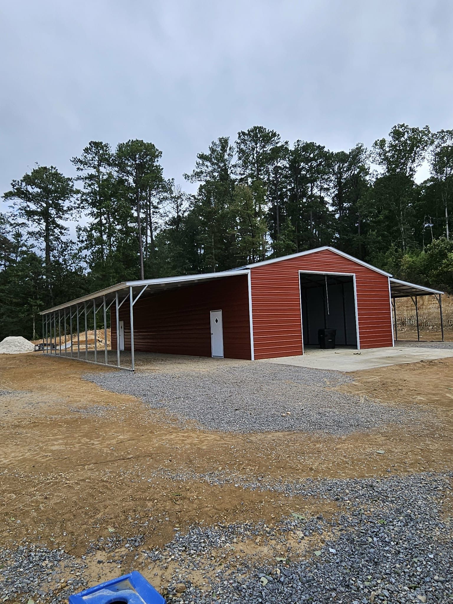 Red metal barn-like shed on a gravel lot with pine trees in the background.