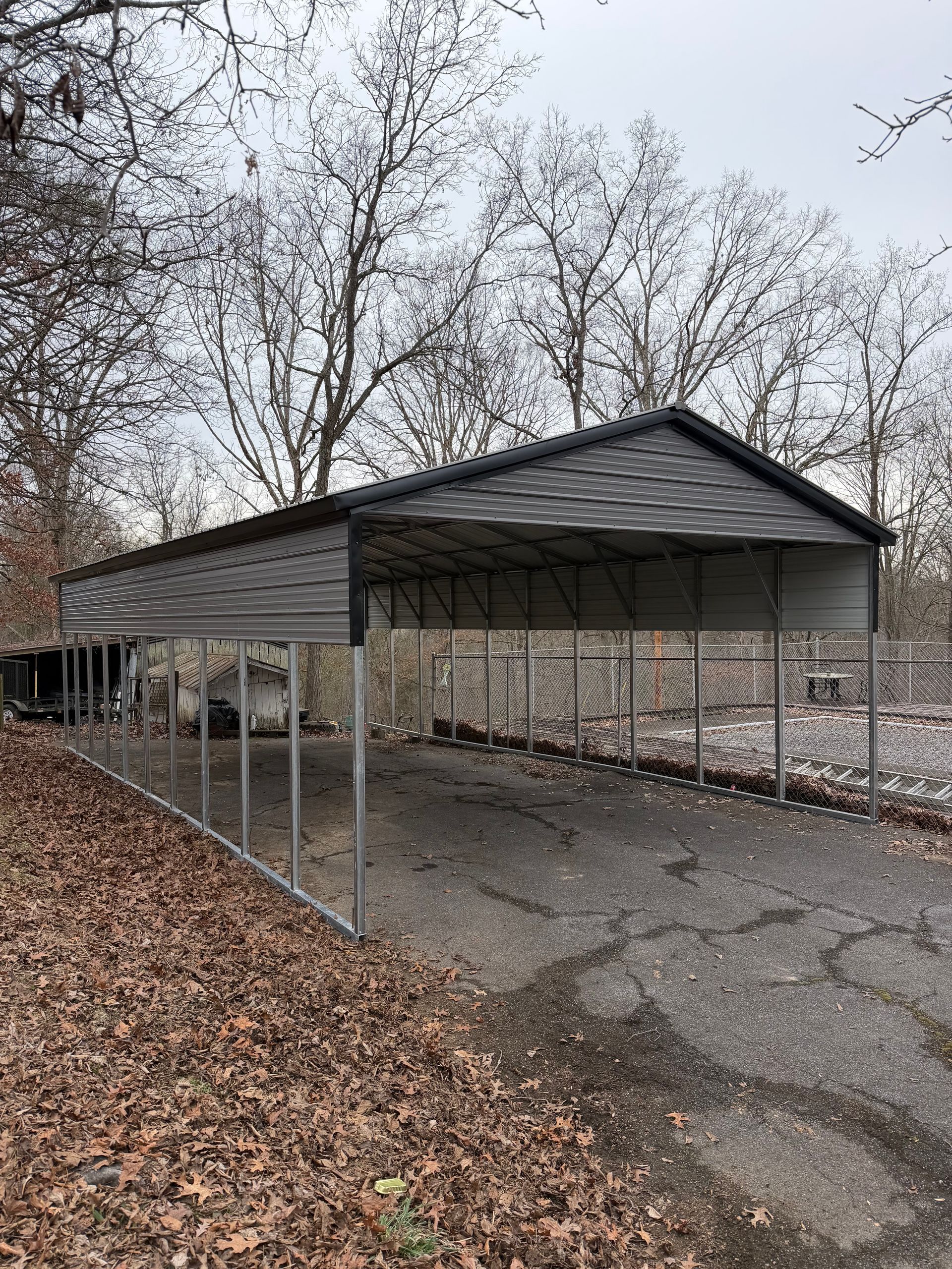 Long metal carport with a dark roof beside a leaf-covered parking lot in winter.