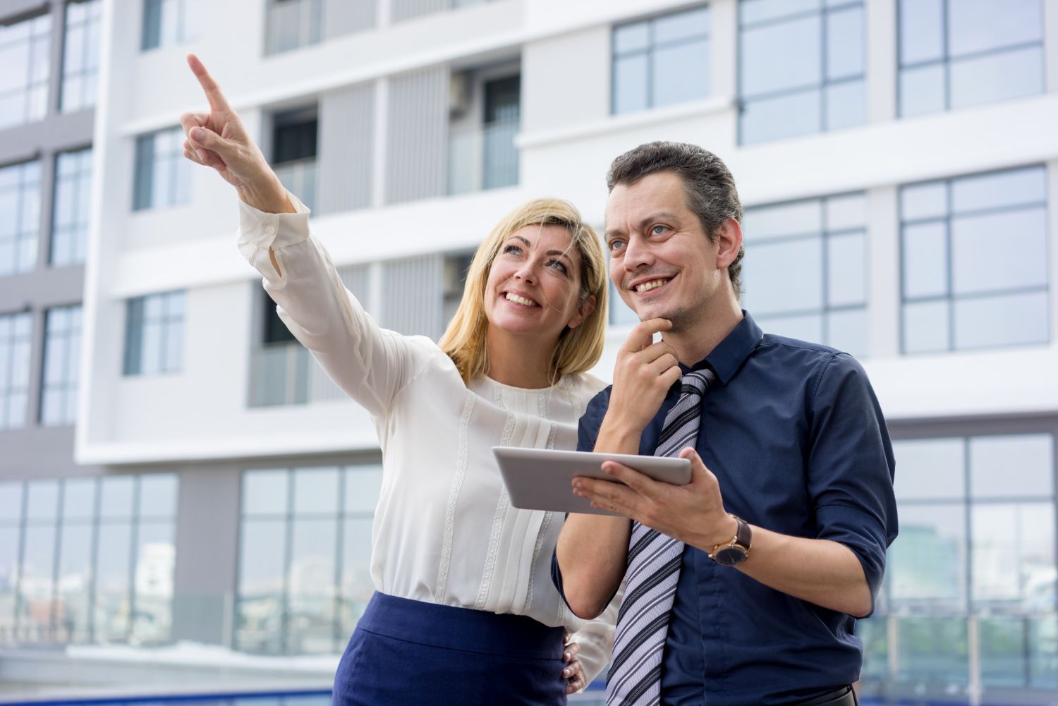 Woman points upward; man holds tablet, both smiling, near modern building.