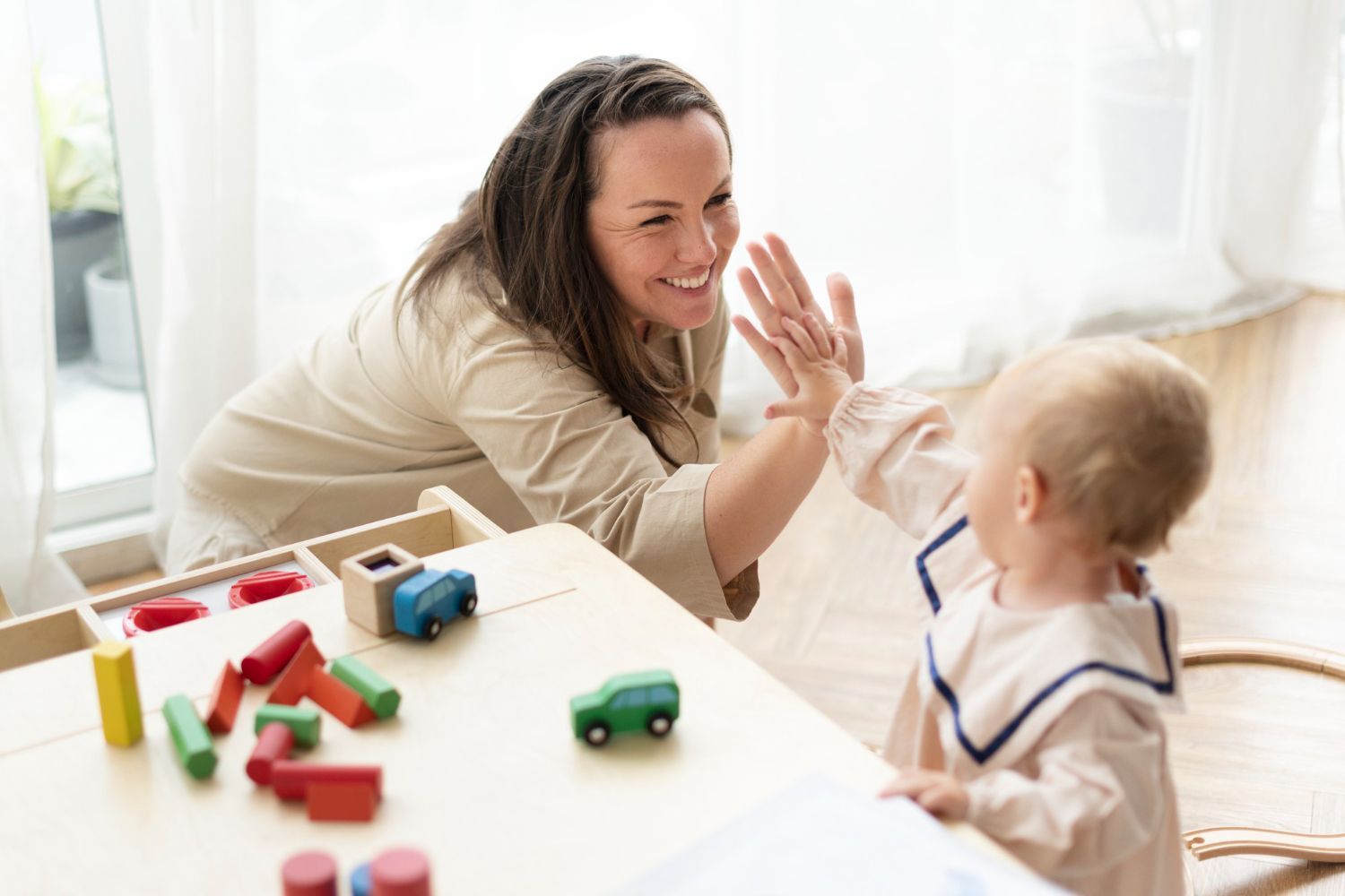 Woman and baby playing, giving high five at a table with colorful wooden toys indoors.