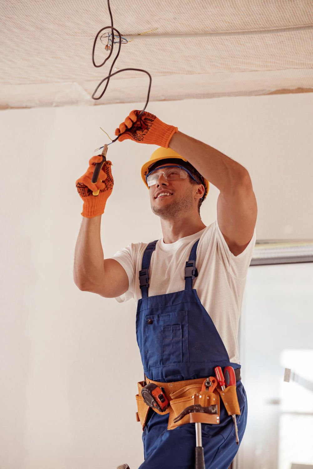 Electrician working on ceiling wires wearing safety gear, in a room.