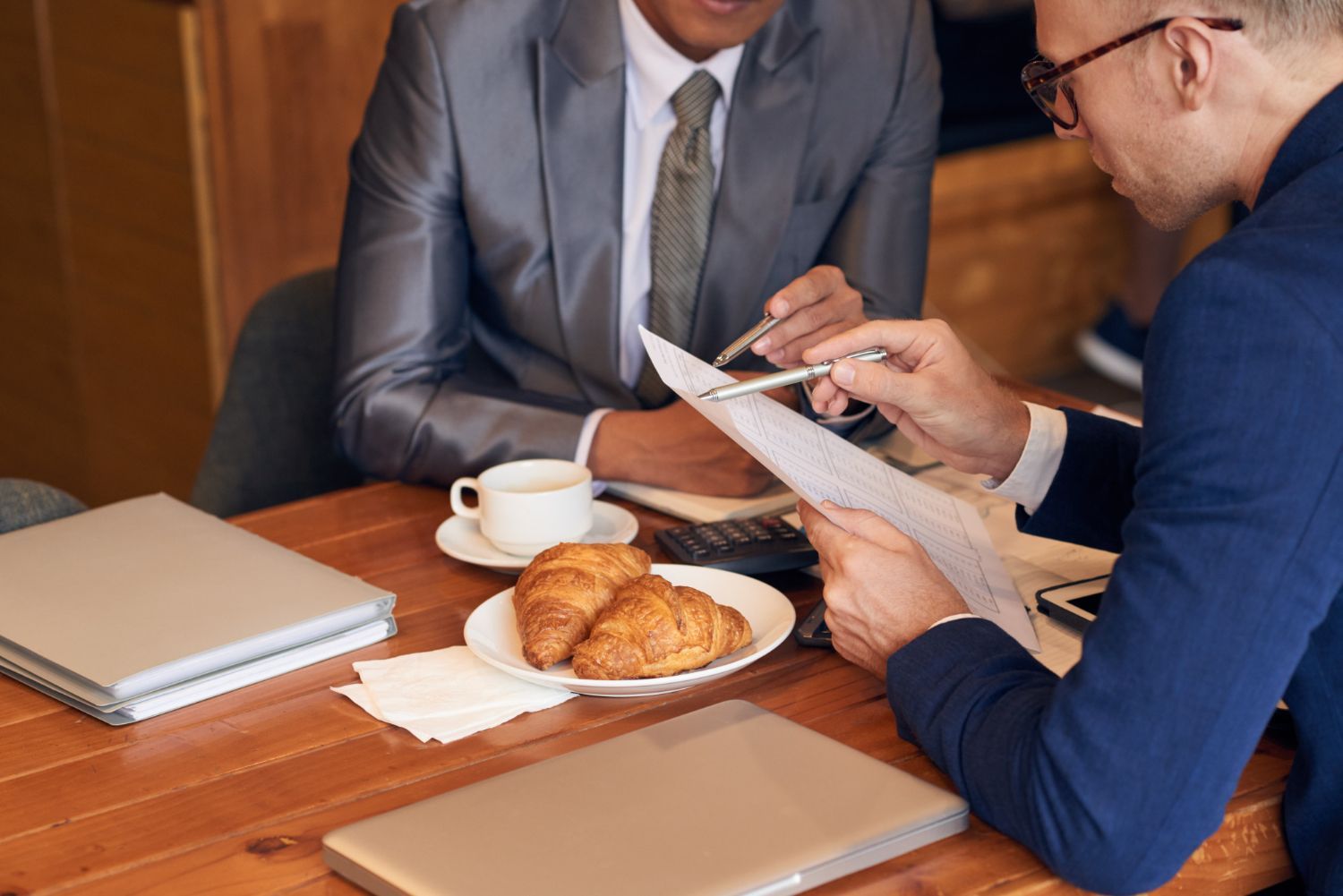 Two men in suits reviewing documents at a table; a coffee cup and croissants are nearby.