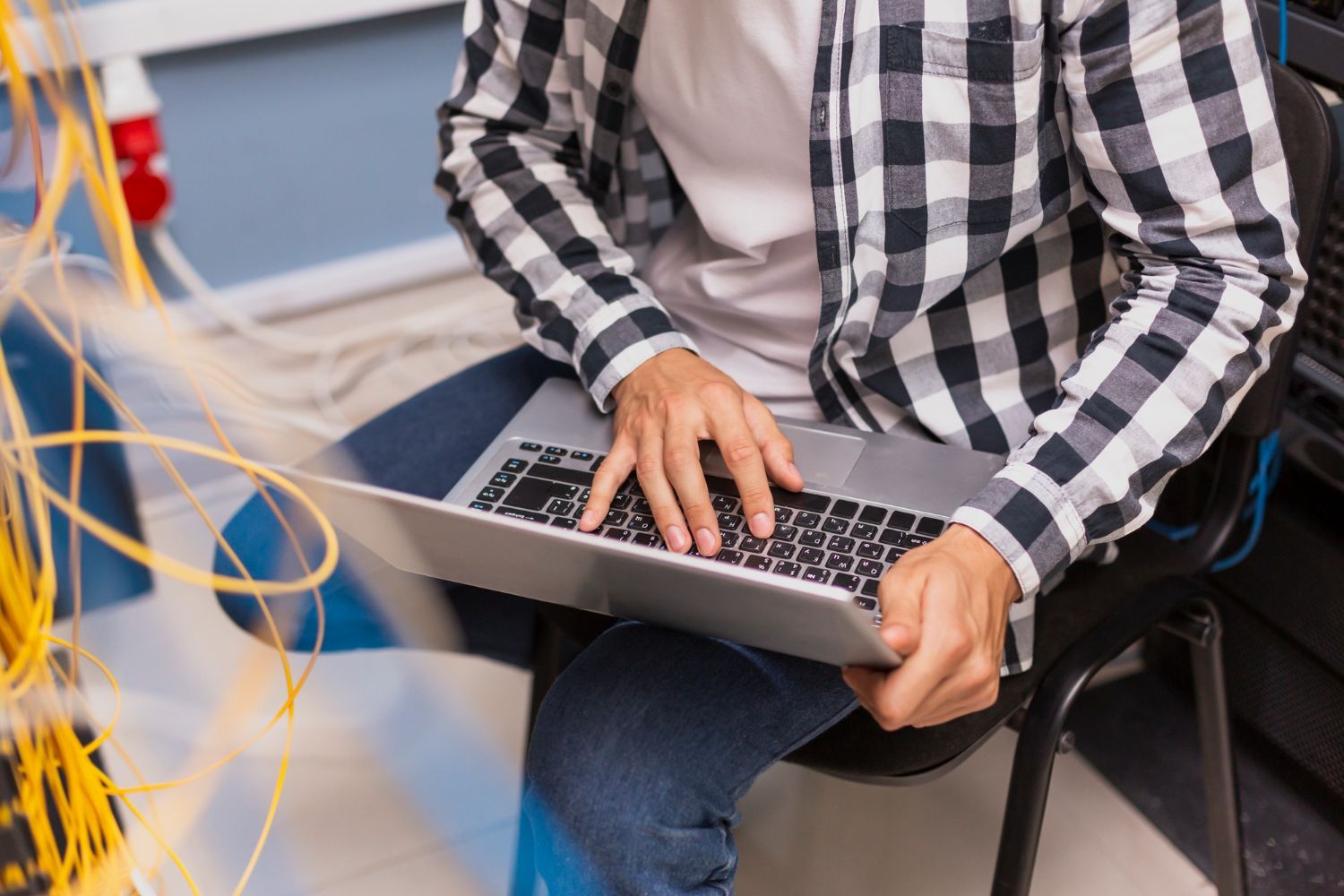 Person with a laptop, troubleshooting network cables in a server room, wearing a plaid shirt.