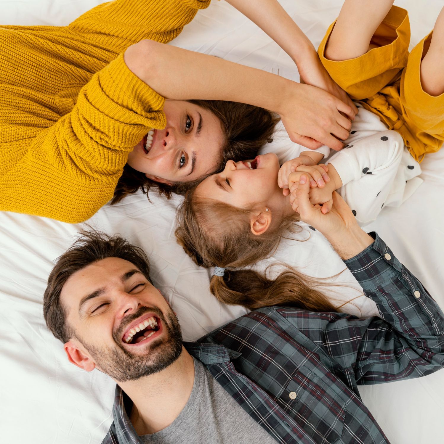 Family of three laughing and playing together on a white bed.