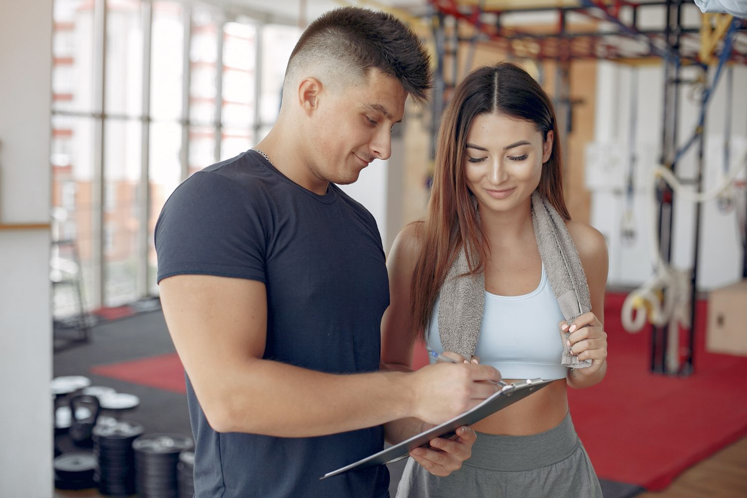 Man and woman in gym looking at clipboard; man points, woman smiles.