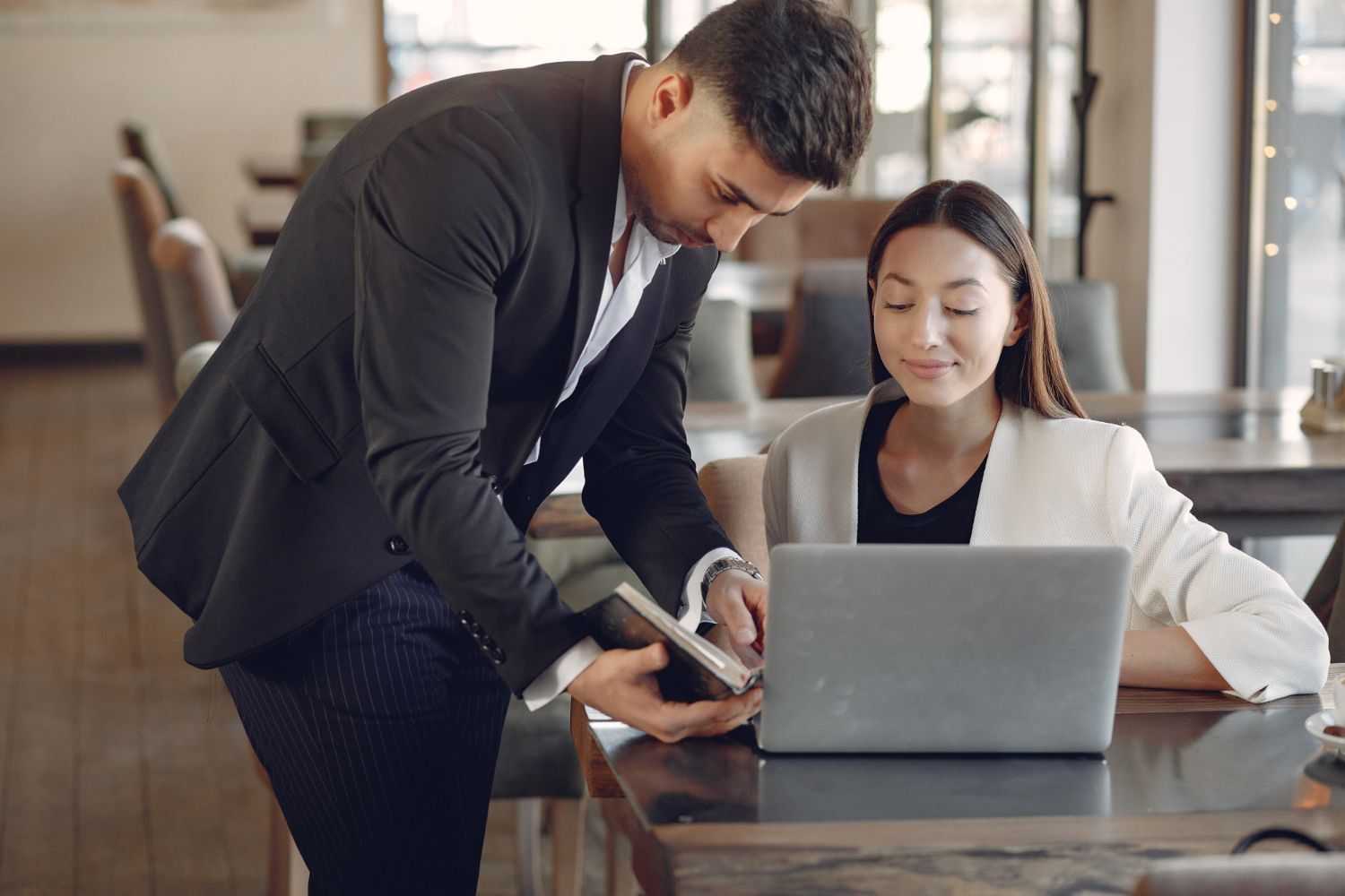Man leans over woman at laptop, showing something on a tablet. Indoors, cafe setting.