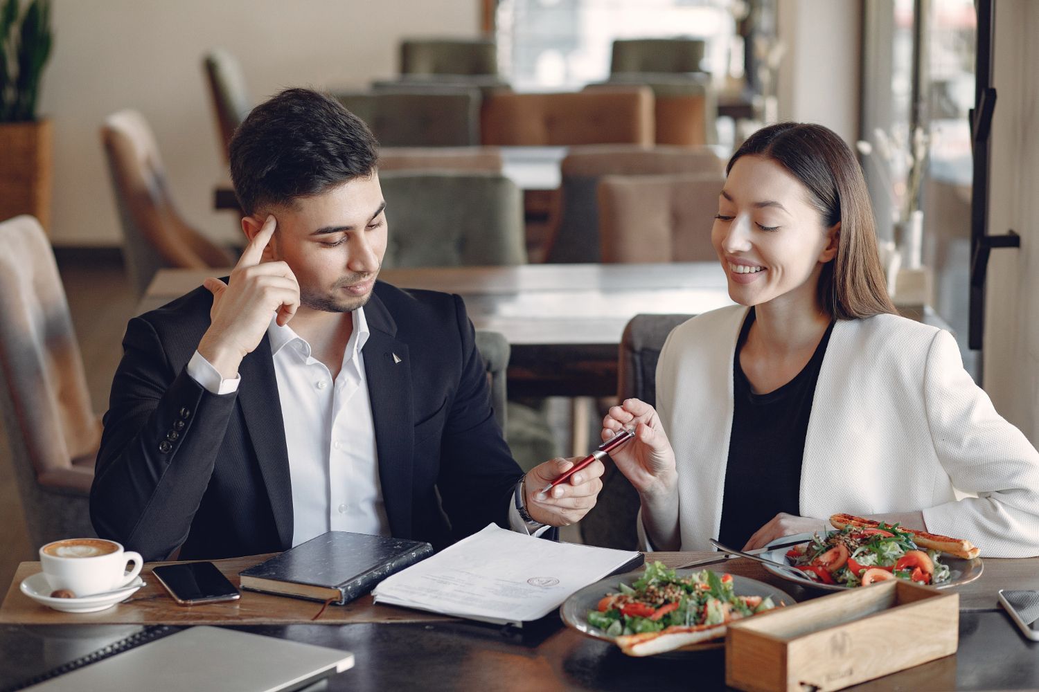 Man and woman in a restaurant, reviewing documents. Woman smiles, holding a pen. Man has hand on head.