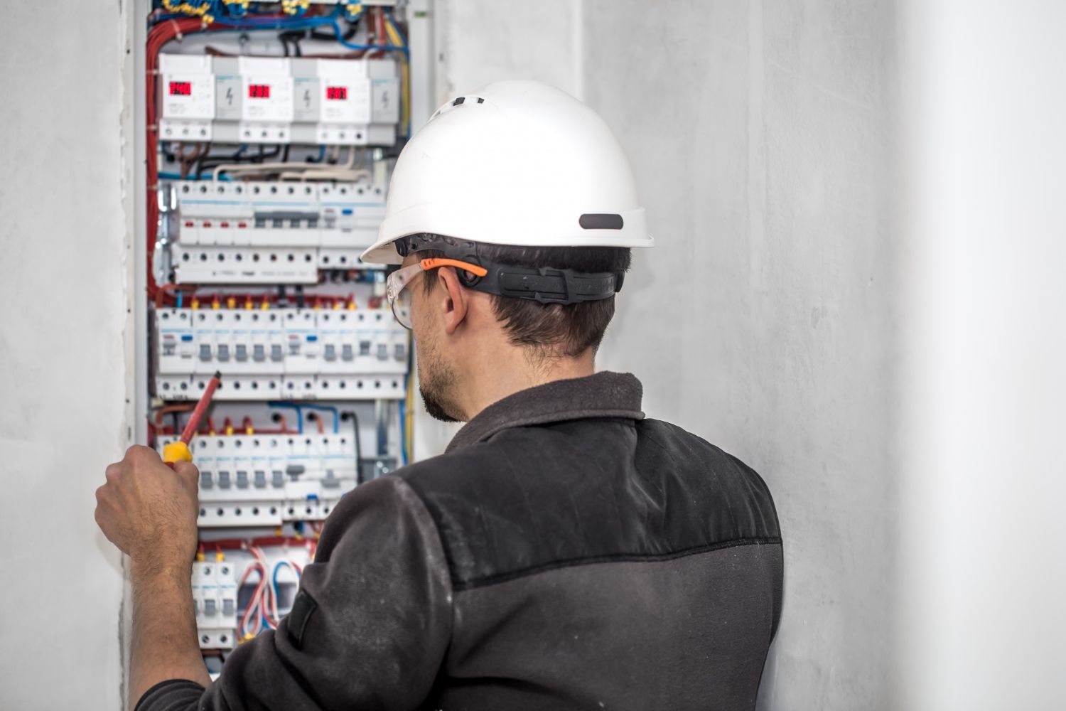 Electrician in a white hard hat and safety glasses working on a circuit breaker panel.