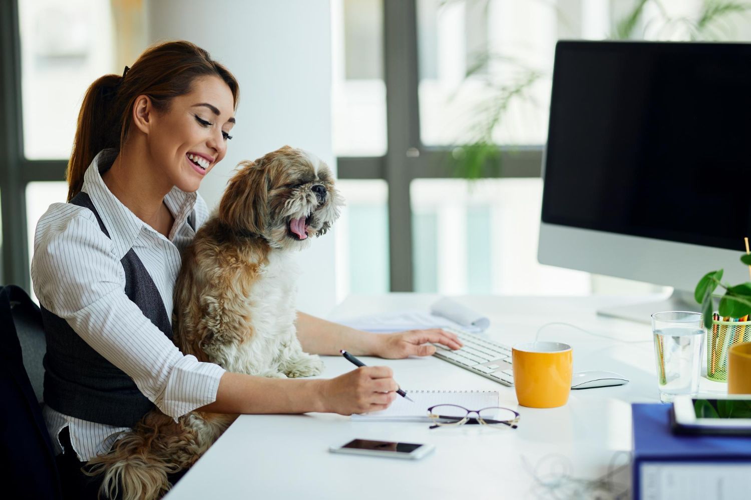 Woman smiles, dog sits on her lap at a desk, writing in a notebook, computer nearby.