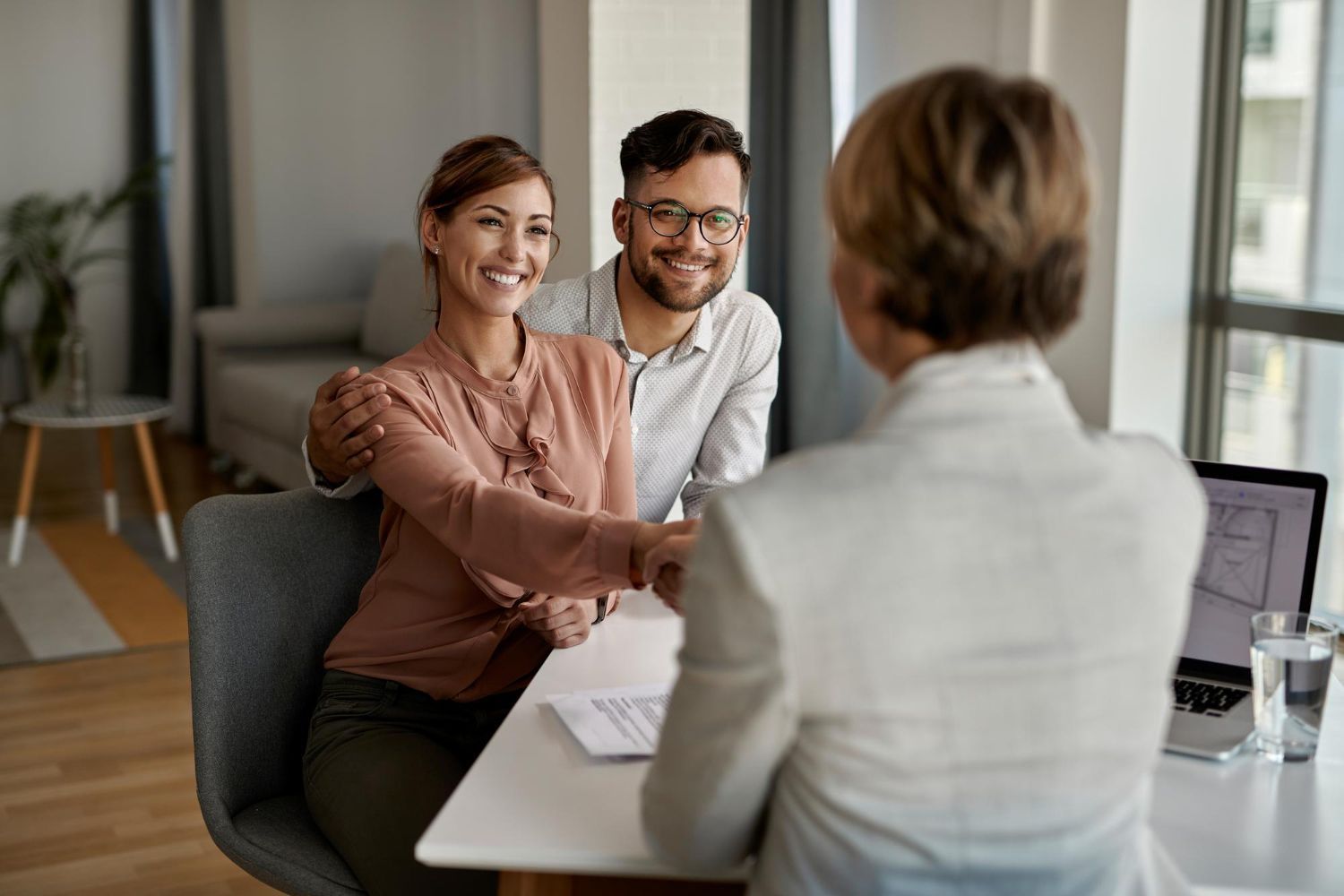 Couple shaking hands with a person, sitting at a table with documents and a laptop in a bright room.
