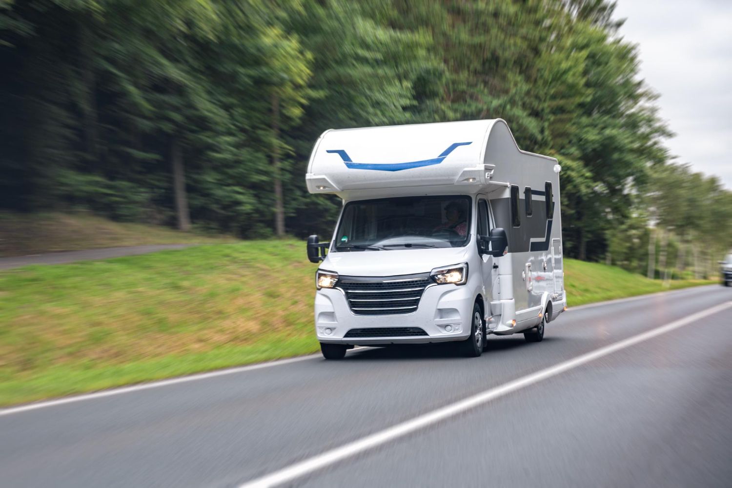 White RV driving on a highway with green trees in the background.