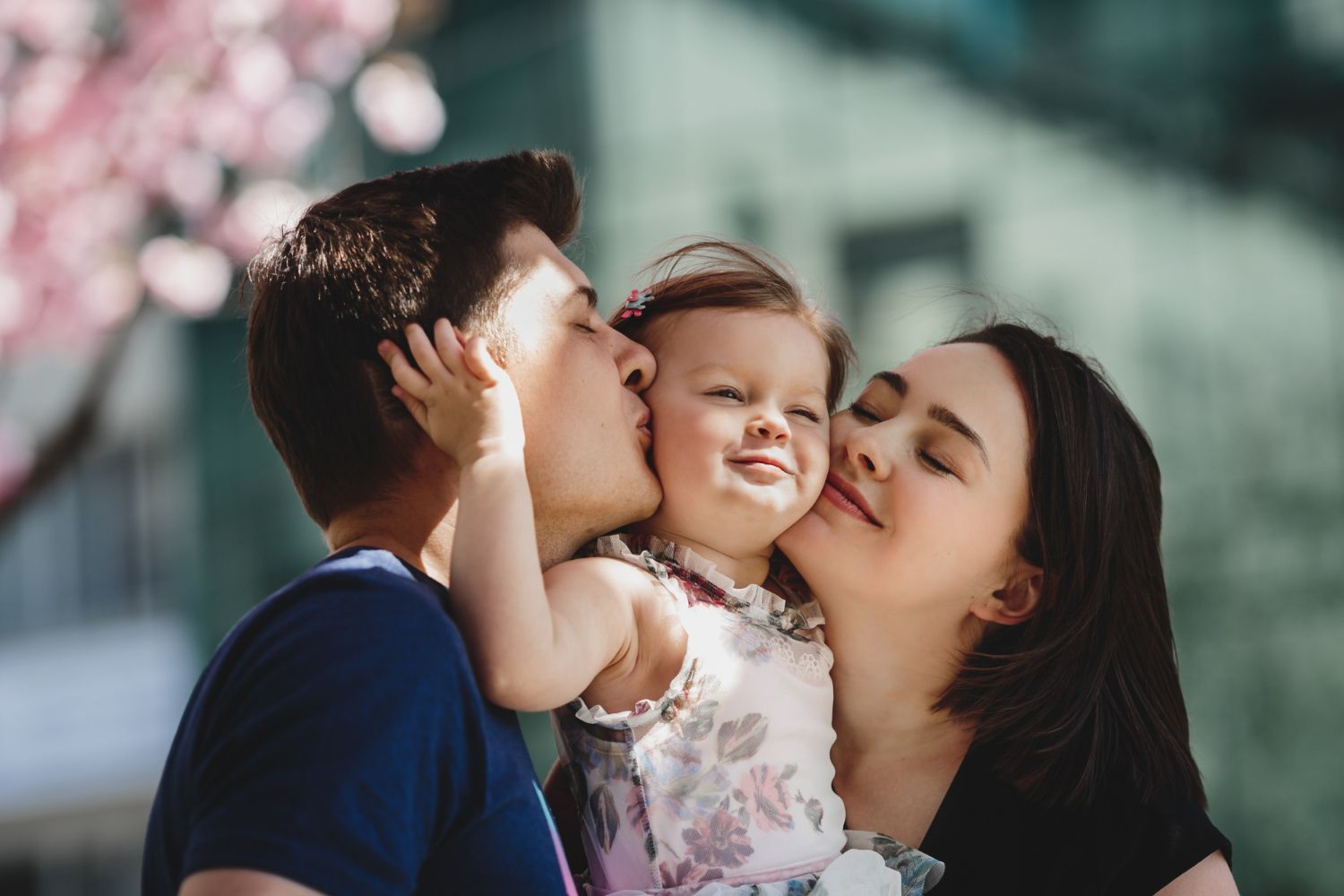 Parents kissing a smiling toddler outdoors near a pink-flowered tree and a building.