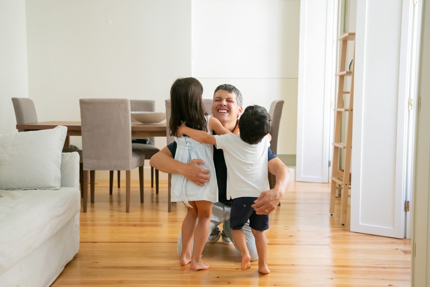 Man kneels, embraced by two children. Indoors, wooden floor, smiles, dining table, light colors.