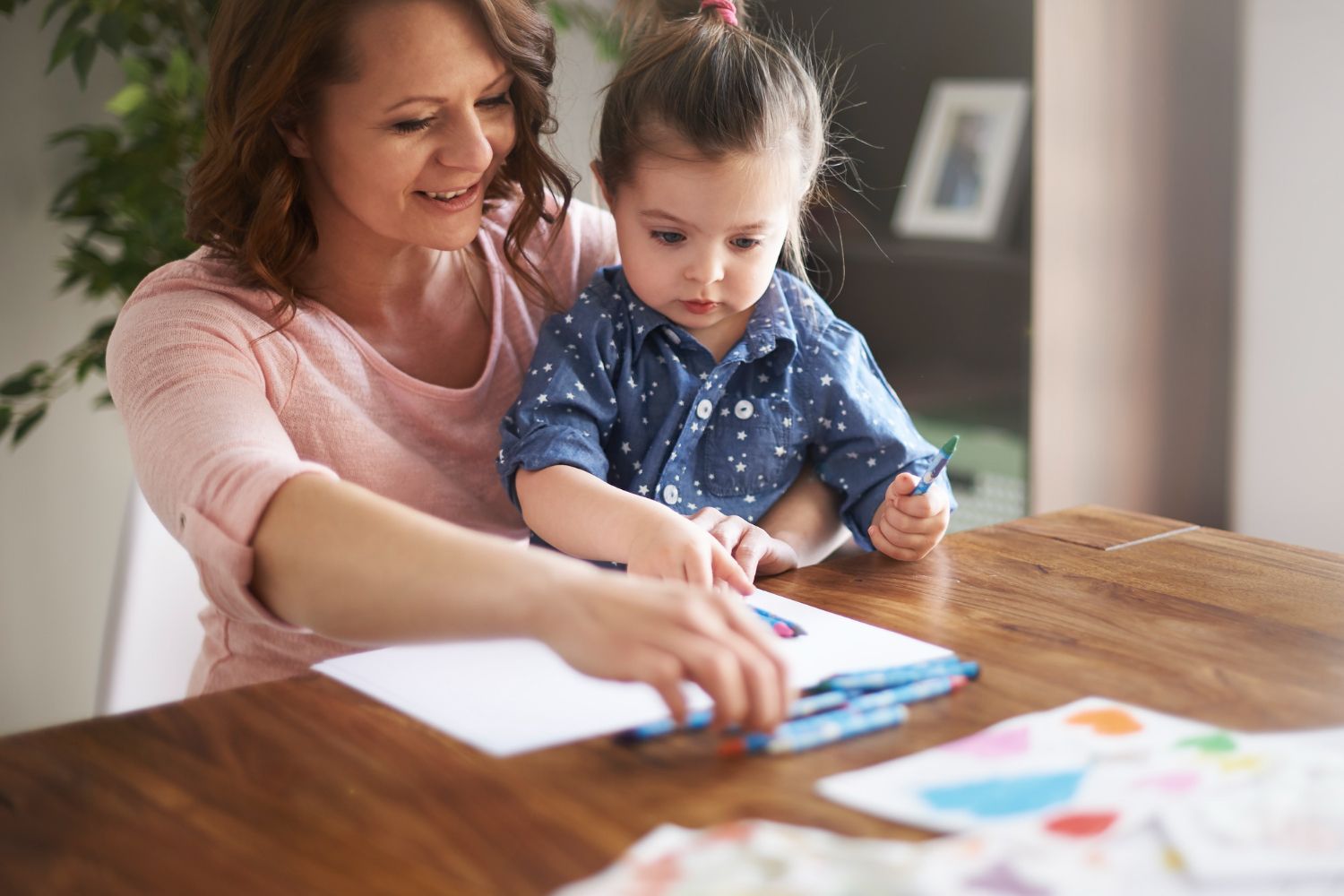 Woman and child drawing together at a table. The child holds a crayon, looking at the paper.