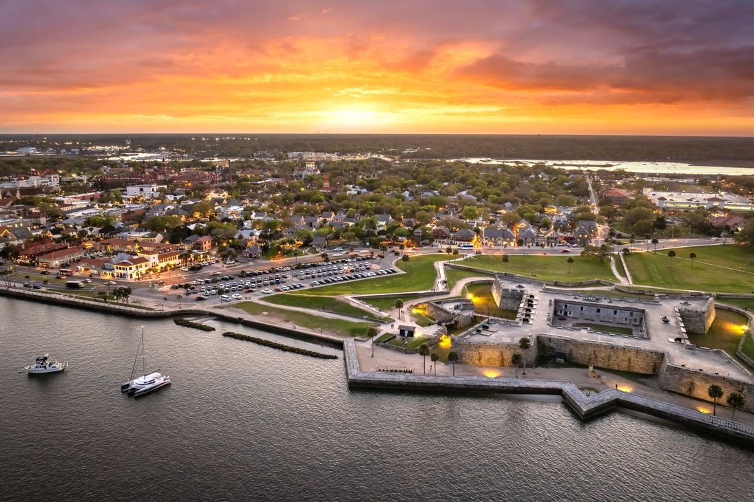 Sunset over a coastal city with a star-shaped fort, boats on the water, and a vibrant orange sky.