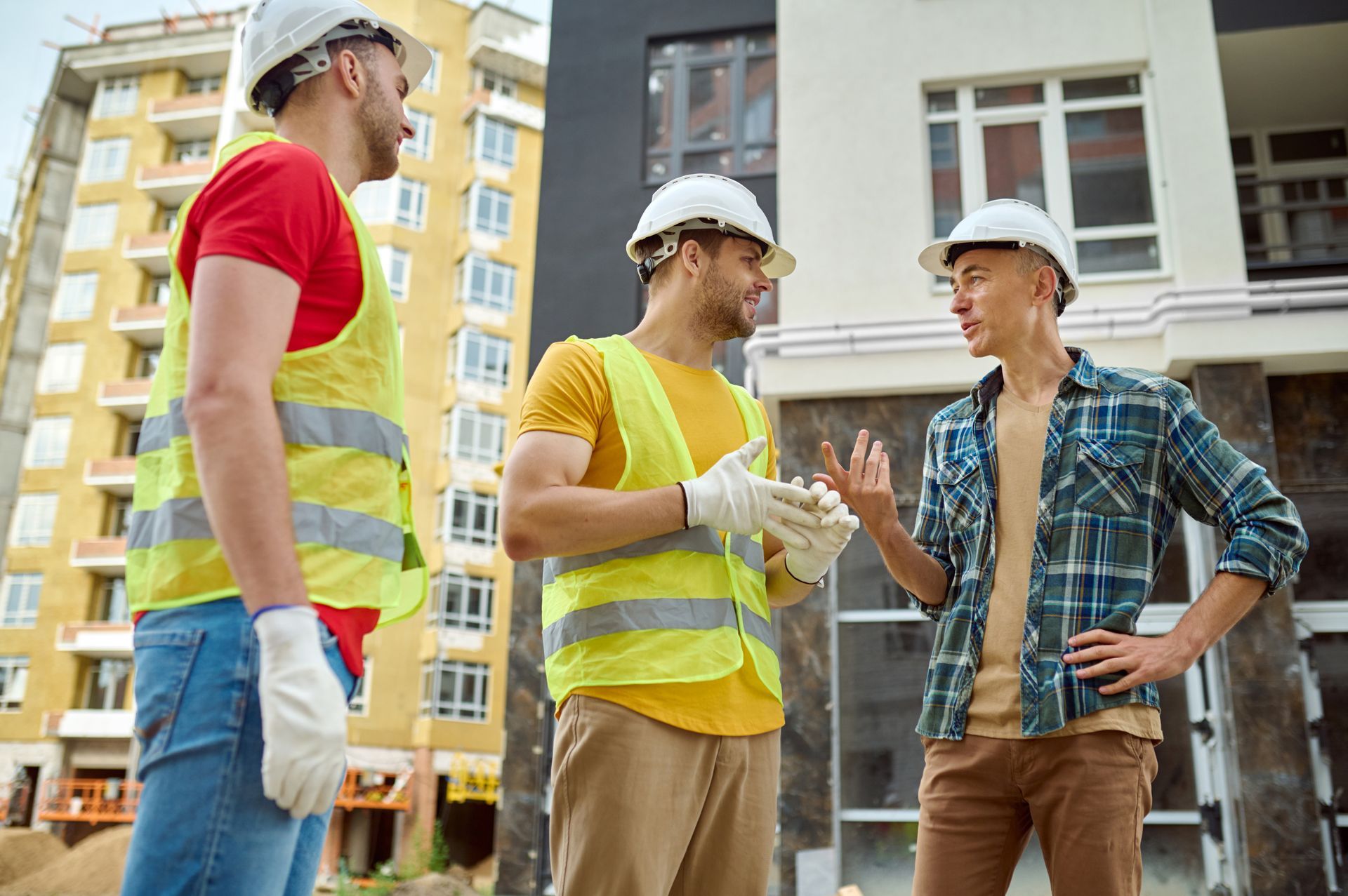 Three construction workers in hard hats and vests conversing outside a building.