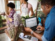 Man working on laptop in flower shop; other family members arrange flowers.