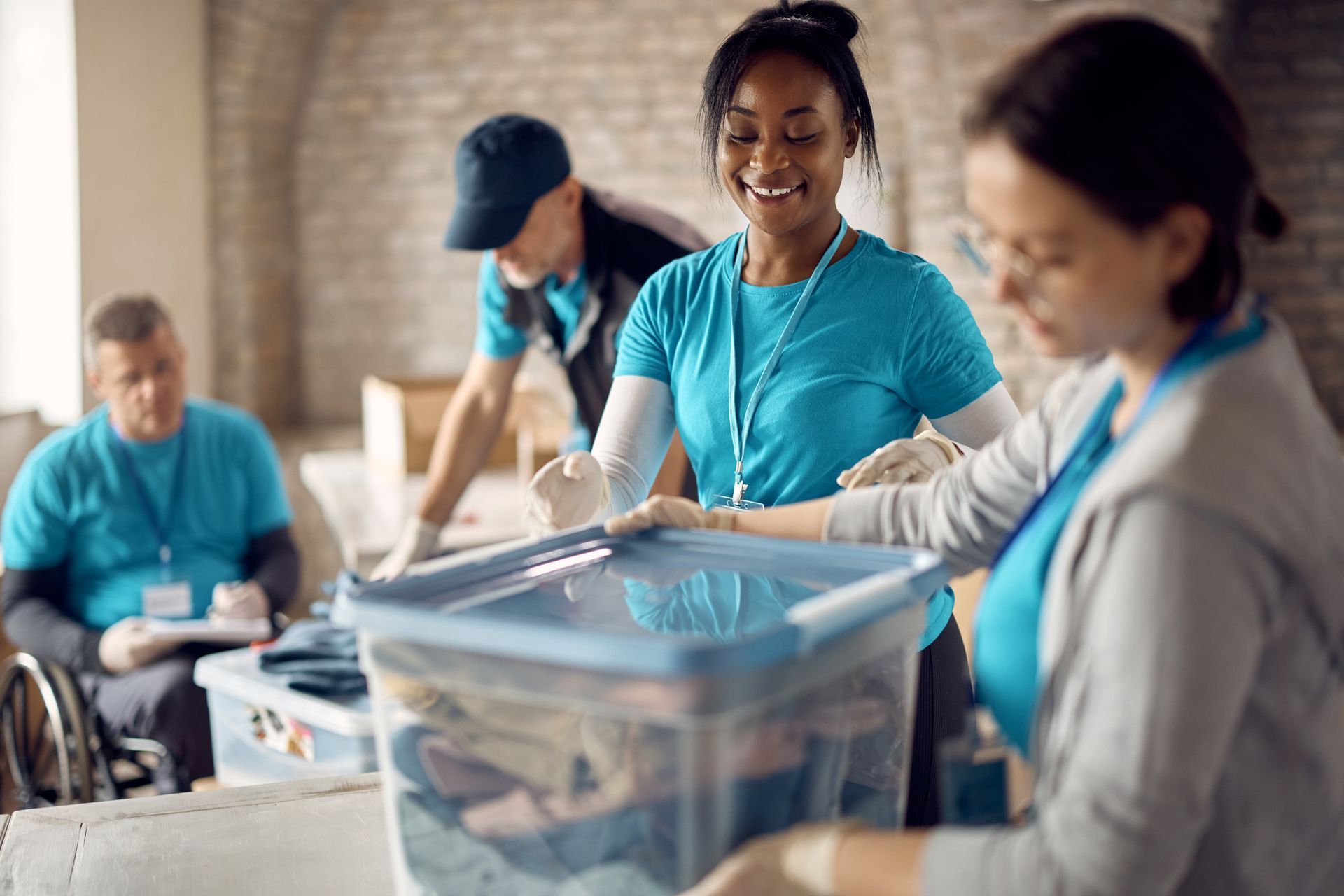 Volunteers sorting donations in a room. Smiling woman holds a bin with others. One man is in a wheelchair.