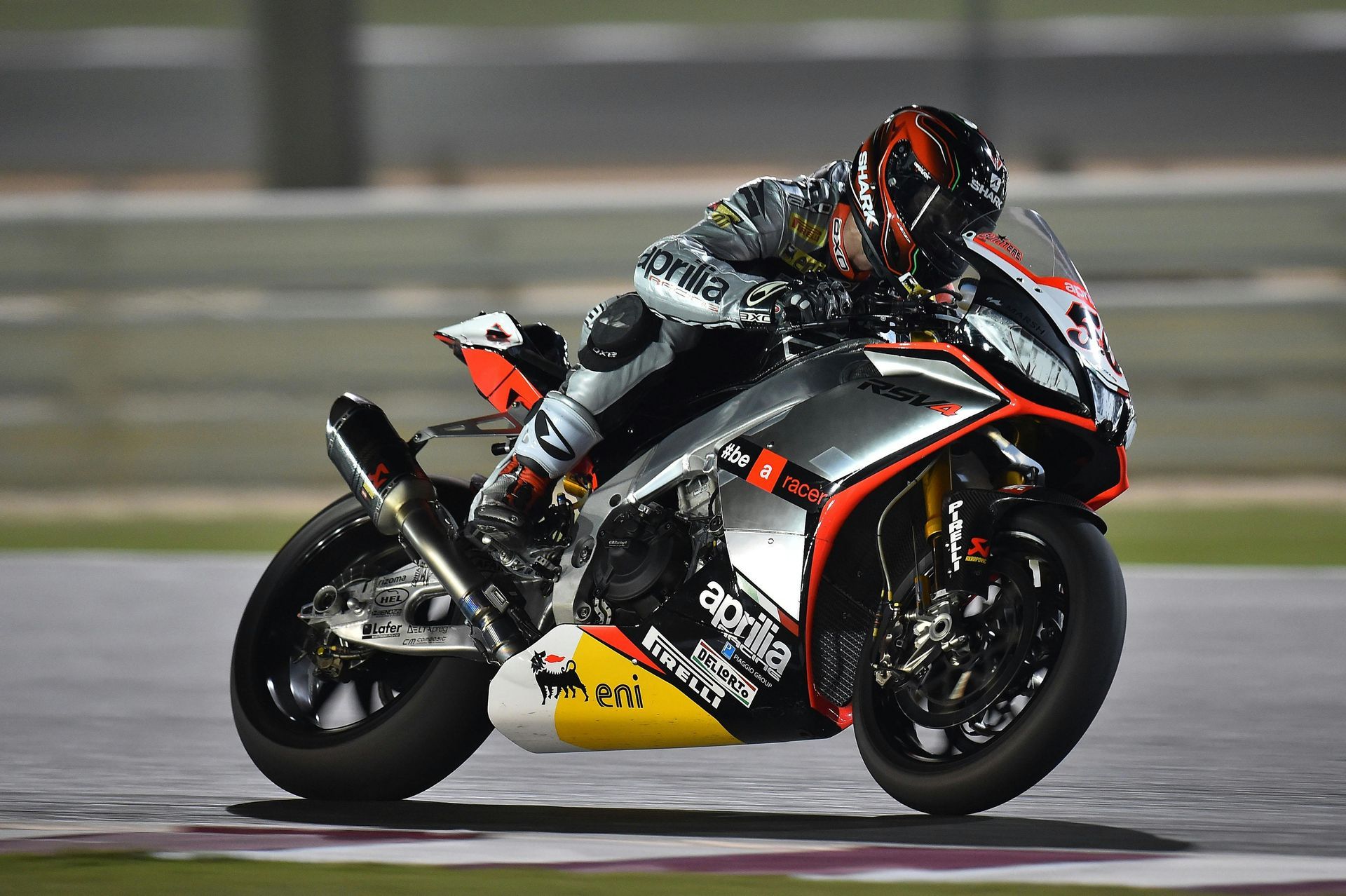 Motorcyclist leans into a turn on a racetrack, Aprilia bike, silver and black colors, night setting.