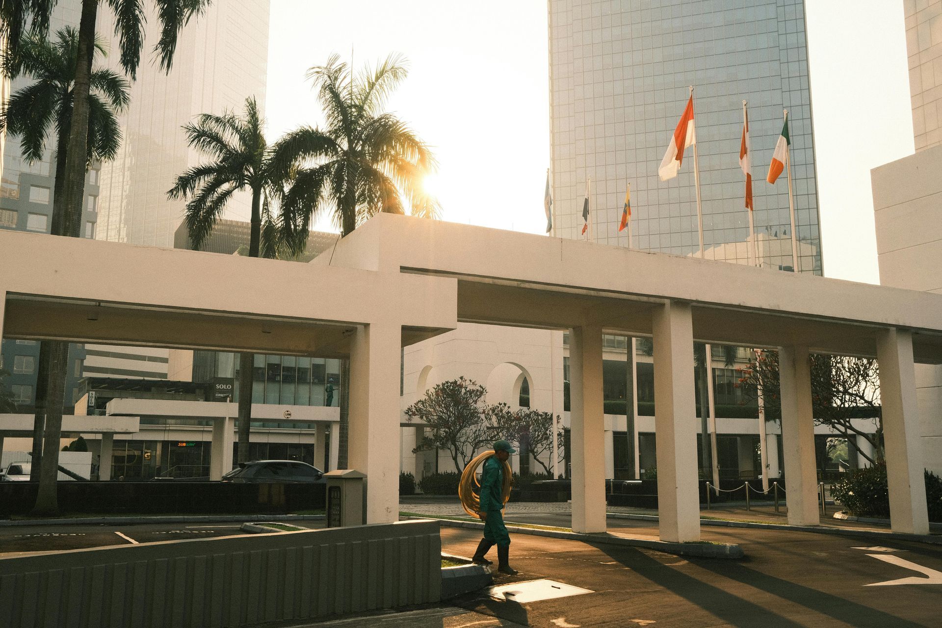 A person in teal walks near a modern building with flags waving in the sun.