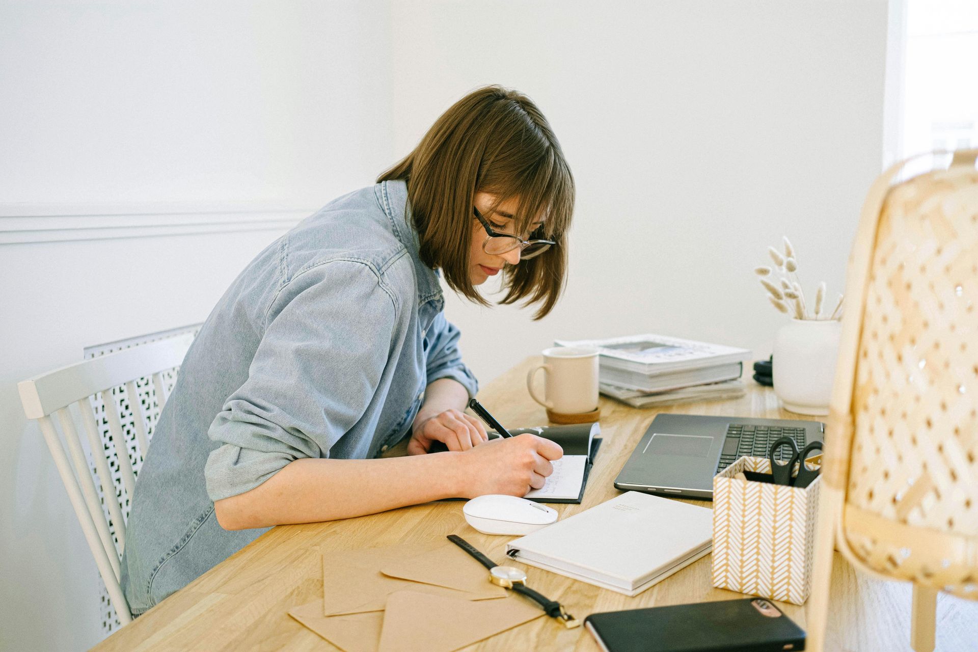 Woman in glasses writing at a desk, sunlight, with laptop and coffee, near a wicker lamp.