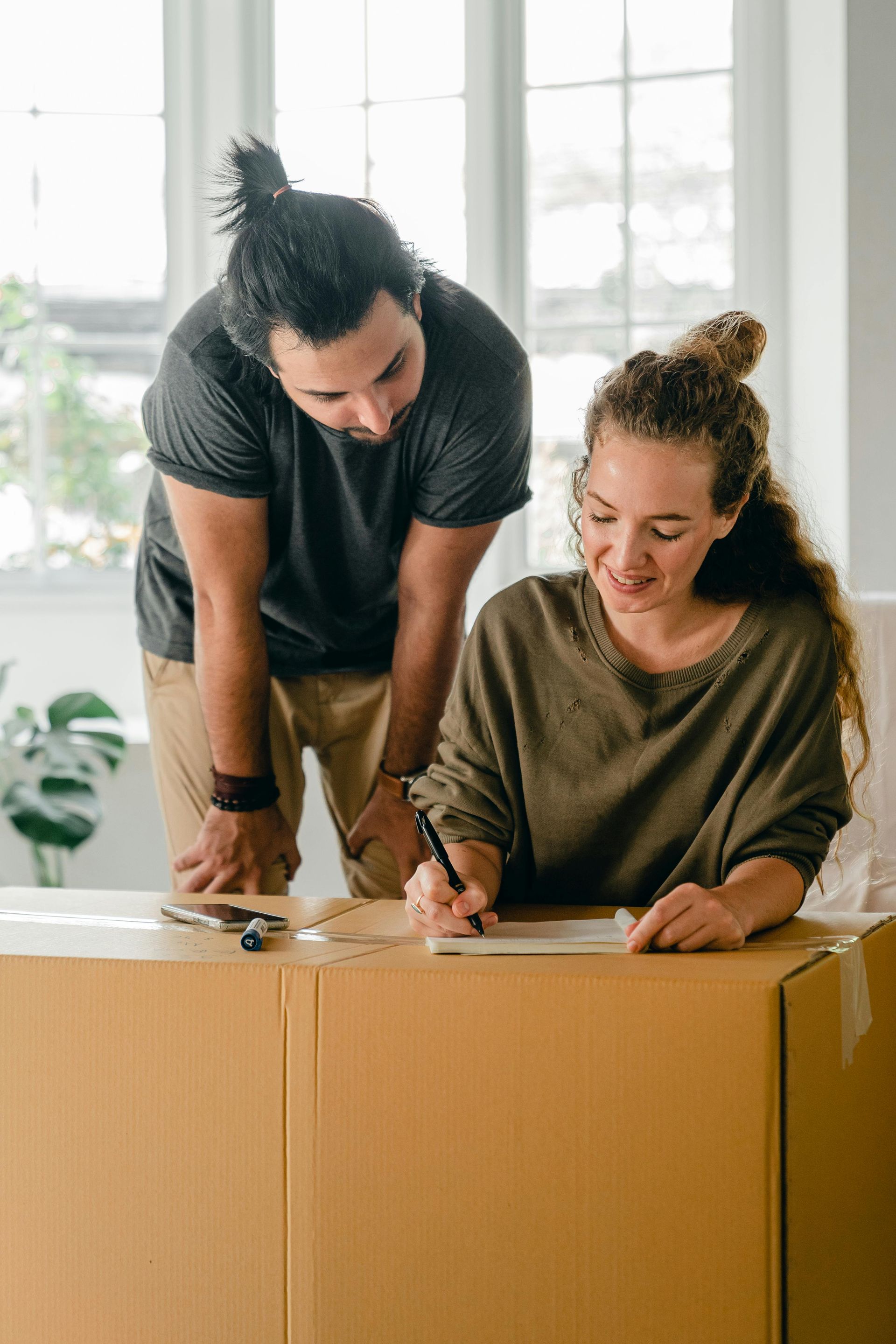 Man leans over woman writing on a box; they appear to be moving into a new home.