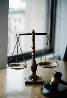 Scales of justice on wooden table next to a gavel and a window.