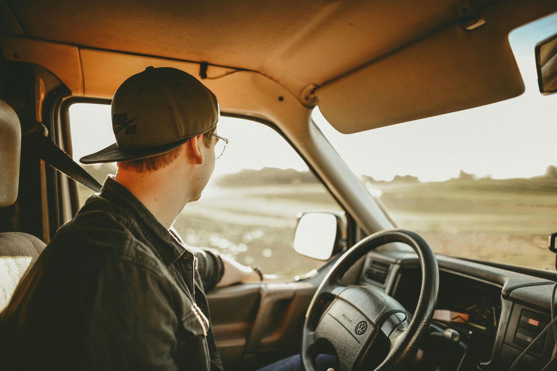Person wearing a hat and glasses driving a vehicle, looking towards the sunlit road.