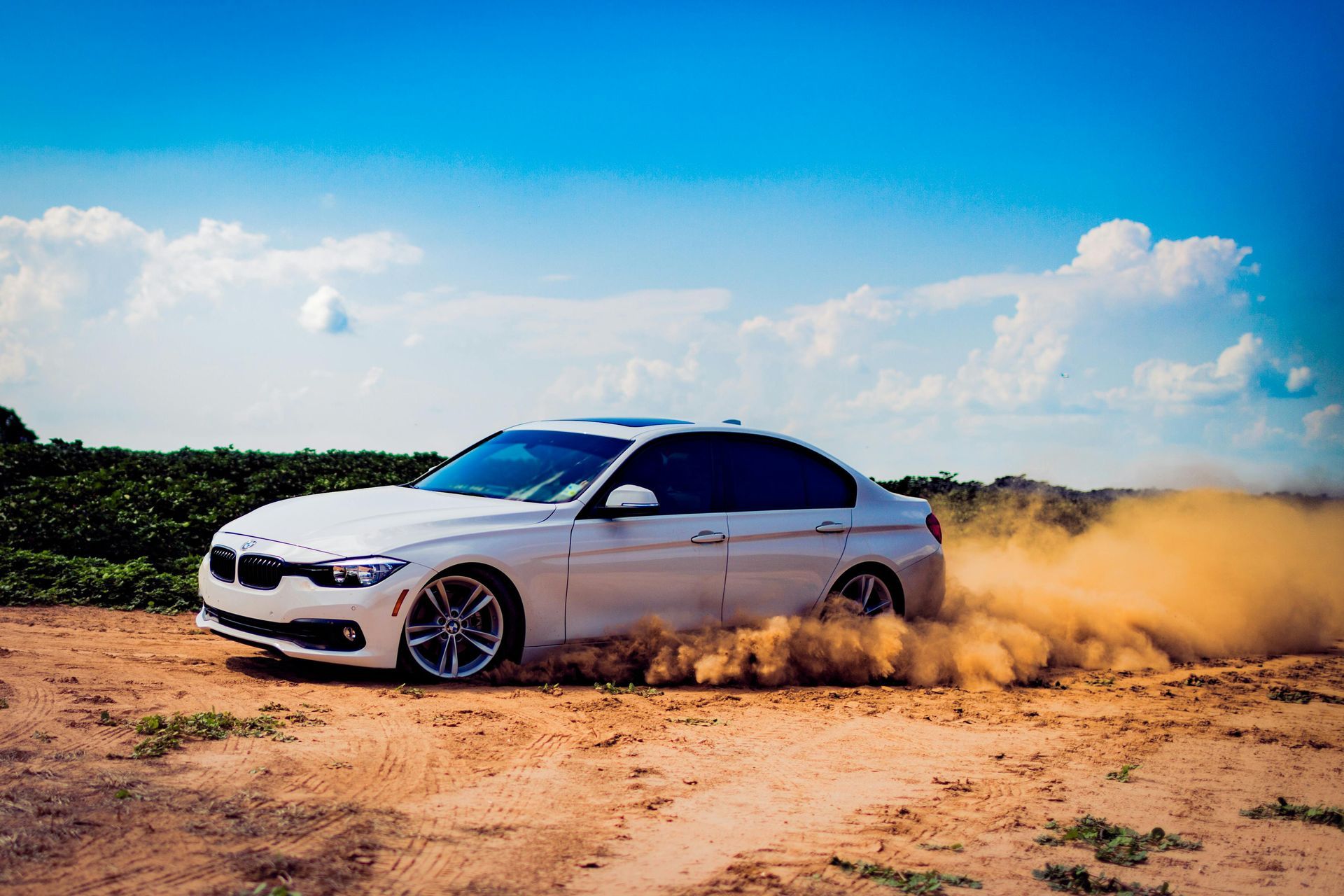 White car drifting on dirt road, kicking up dust cloud under a blue sky.